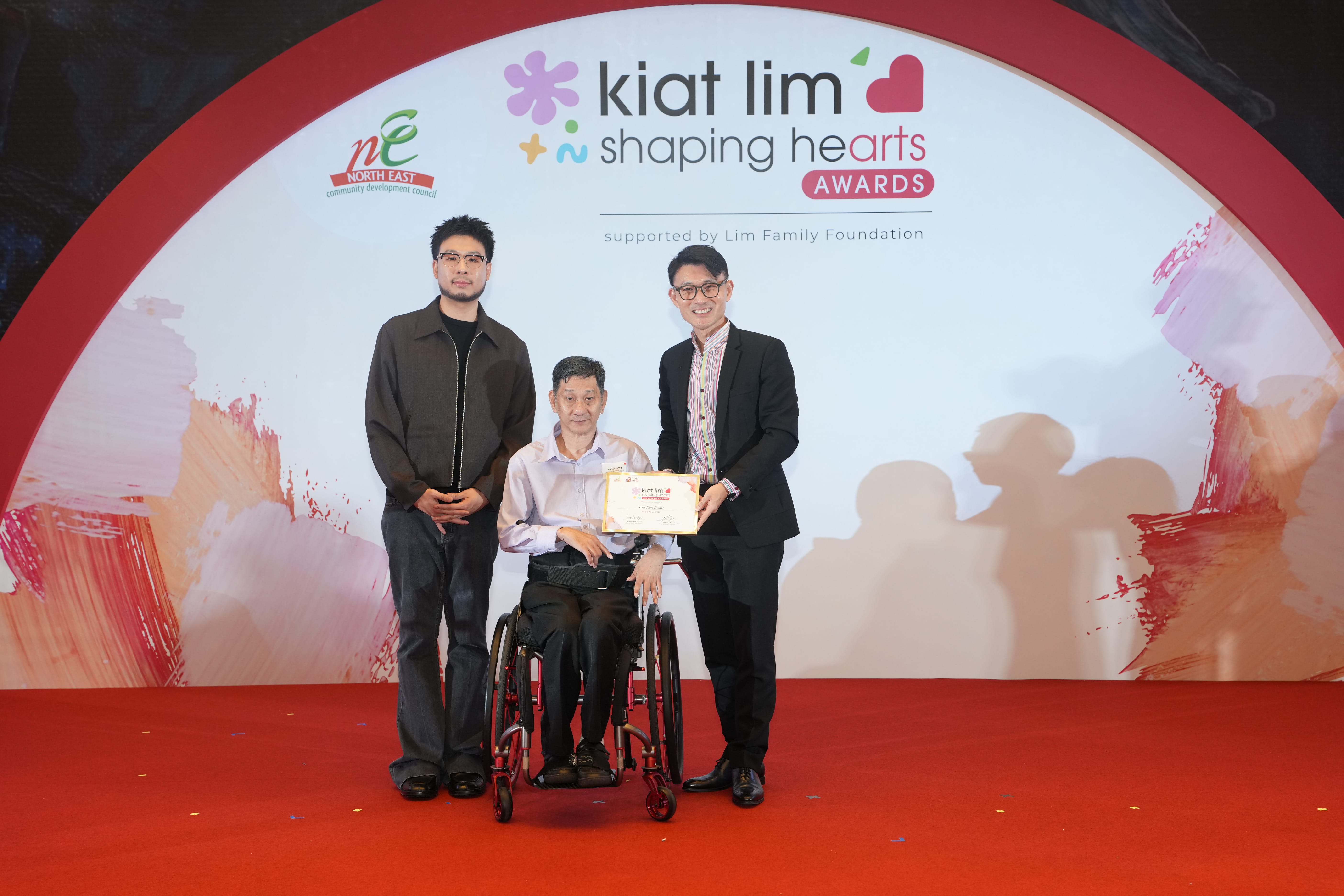 Award recipient standing on stage with the presenter, holding their certificate and posing for a group photo during the award ceremony, with a backdrop reading ‘Kiat Lim-Shaping Hearts Awards'.